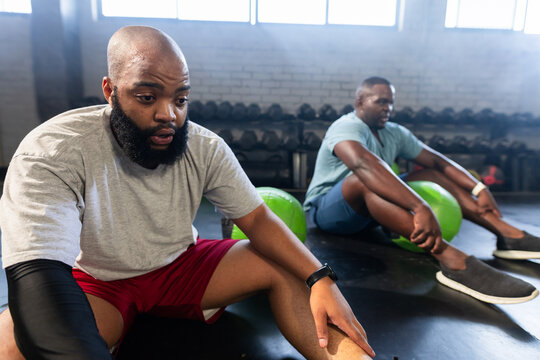 African male workout partners resting and stretching on warehouse gym floor near dumbbell rack