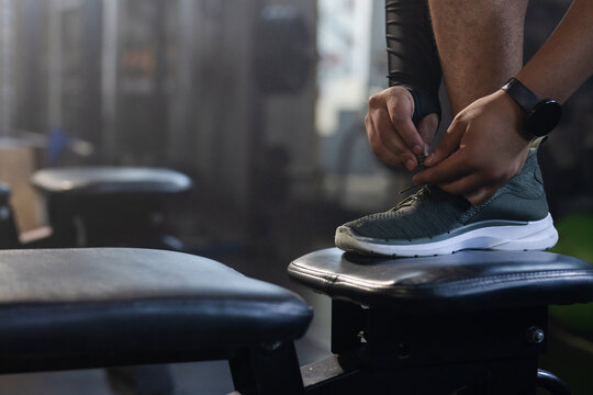 Right foot and hands are tying knit sneaker laces on padded gym bench, smartwatch visible