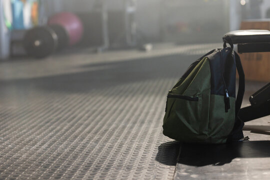 Olive-green backpack is resting on textured rubber gym floor near bench leg, blurred weights behind