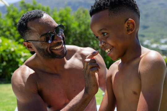 African American shirtless dad wearing round shades putting sunscreen on boy with finger on lawn