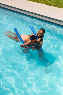 African American father holding child using blue pool noodles in backyard pool with orange swimsuit