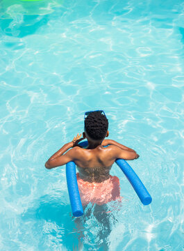 African teen standing waist-high in turquoise pool holding blue noodle, in pink trunks and goggles