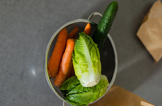 Metal colander holding three carrots, romaine and cucumber on dark gray countertop near paper bag