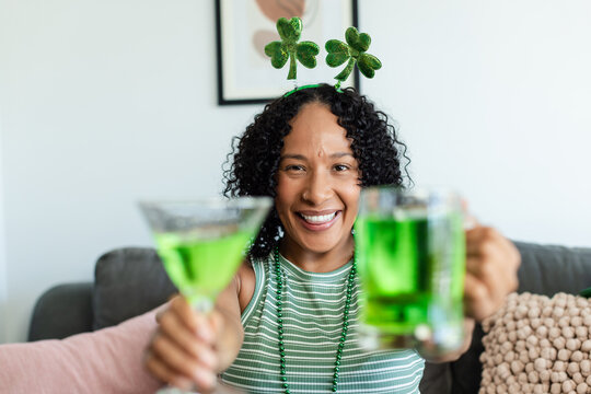 Adult African woman sitting on gray sofa holding two green drinks wearing shamrock headband smiling