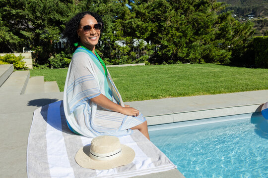 African American woman sitting on striped towel at pool edge wearing sunglasses and straw sun hat
