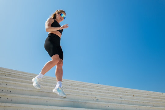 Woman exercises on stairs under clear blue sky during daytime while wearing sports gear and sunglasses