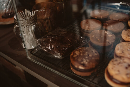 Close-up of assorted homemade cookies arranged on a metal rack inside a glass bakery display case. Warm ambient lighting, shallow depth of field, and cozy atmosphere create a rustic cafe vibe. 