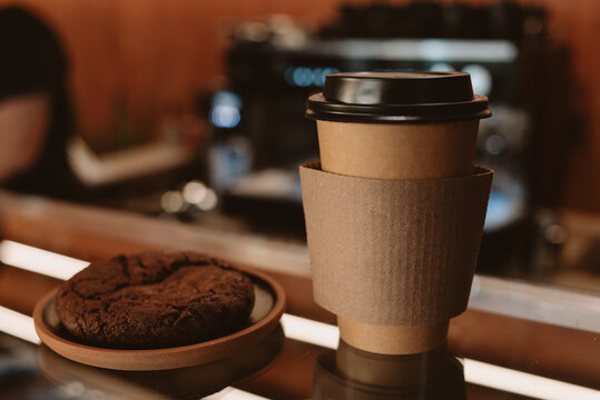 Close-up of a takeaway coffee cup with kraft sleeve and lid placed on a coffee counter beside a chocolate cookie. Warm ambient lighting and shallow depth of field create a cozy coffee shop atmosphere,