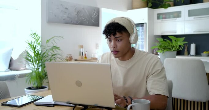 African male adult starting work, sitting in kitchen, typing on laptop then stretching, refocusing
