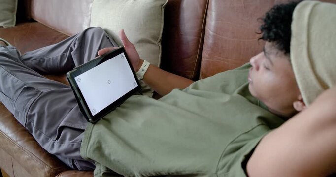 Teen male reclining on brown leather couch is seeing blank tablet screen and reaching, reading