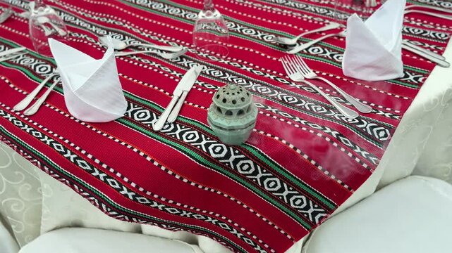 Close-up of traditional dining table setup with red patterned tablecloth, folded white napkins, cutlery and decorative Ramadan lantern, prepared for festive meal.