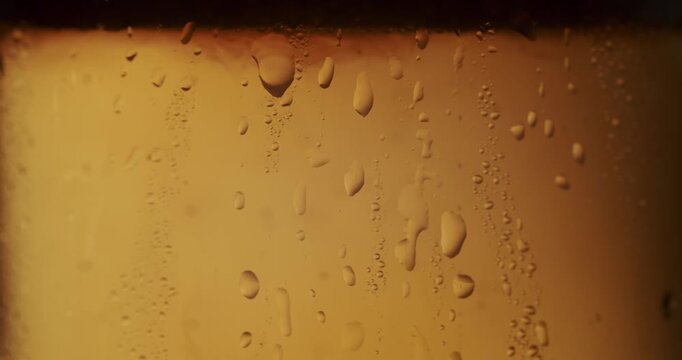 Extreme close-up of condensation water drops sliding down the side of a glass of cold beer in slow motion, creating a refreshing and thirst-quenching visual on a warm golden background