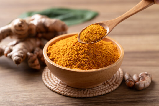 Turmeric powder in a wooden bowl with a wooden spoon scooping it, and fresh turmeric roots on a wooden background. Organic healthy spice