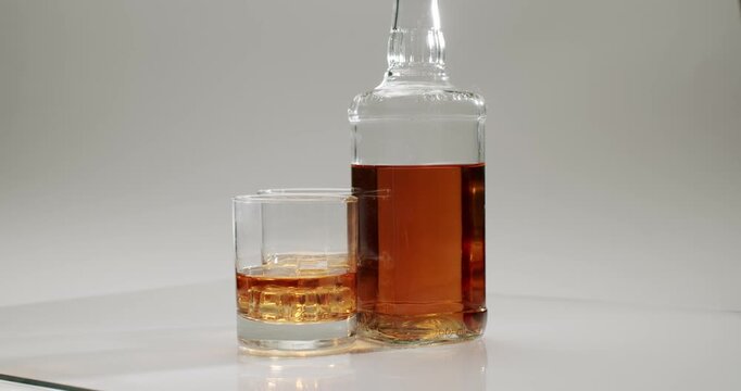 Elegant bottle and a tumbler glass filled with golden brown whiskey and ice cubes spinning in slow motion on a reflective white surface, illustrating luxury, celebration, and relaxation.