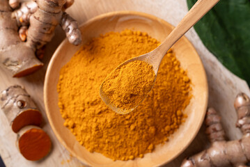 Turmeric powder in a wooden bowl with a wooden spoon scooping it, and fresh turmeric roots on a...