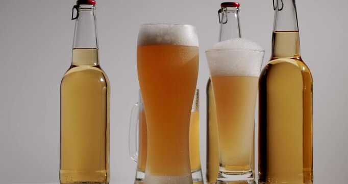 Assorted beer glasses filling with golden lager, creating a thick foam head, surrounded by bottles on a white background. Full bottles and glasses of cold, fresh beer with foam rotating slowly shot.
