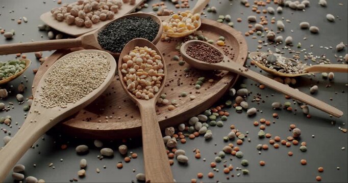 Rich assortment of raw cereals, grains, and legumes, including quinoa, chickpeas, lentils, and sesame seeds, arranged in wooden spoons on a dark surface in a captivating slow-motion display.