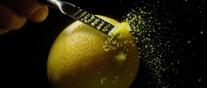 Citrus Zest Spark. Plant-Based & Sustainable Food Preparation. Sensory / Prep. A microplane zesting a bright yellow organic lemon, set against a dark, with particles of zest flying into the air
