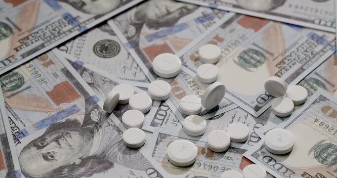 White prescription pills falling in slow motion over a background of scattered one hundred dollar bills, symbolizing the high cost of healthcare, medicine, and the pharmaceutical industry