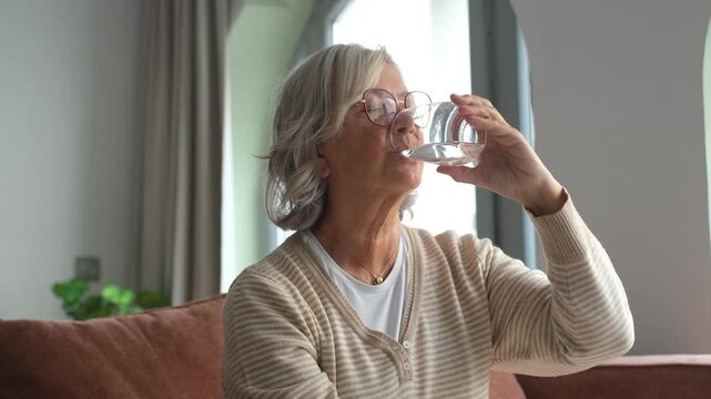 Thirsty elderly woman drink clear pure mineral water from glass, take daily dose for body balance and refreshment, mature female enjoy clean still aqua, hydration, healthy lifestyle concept
