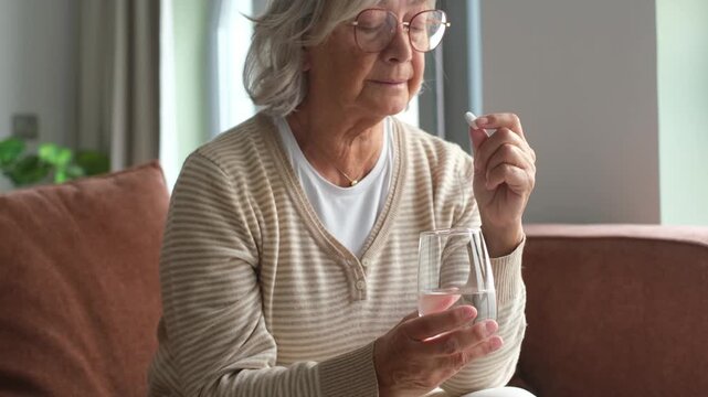 Close up senior mature woman taking painkiller, antibiotic, supplements or vitamin medication, middle aged female holding glass of water and white round pill in hands, disease treatment concept
