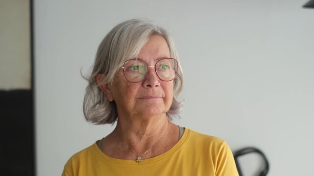 Elderly woman looks out the window with a worried expression. She reflects on her thoughts and feels concerned about her day while standing at home.

