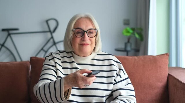 Happy smiling senior woman sitting on couch in the living room, pointing tv remote controller to switch channels, watching favorite movie online series
