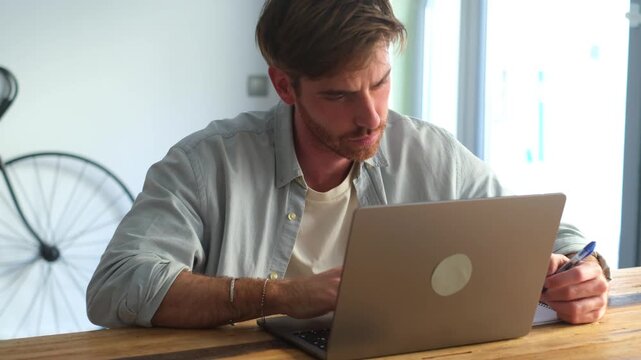 Side view handsome young businessman working with computer remotely, sitting at wooden table in office. Pleasant happy man communicating in social network, searching information online.
