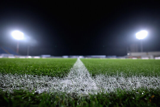 Close-up low-angle view of a white painted line on a green grass sports field at night under bright stadium floodlights. This atmospheric shot captures the essence of competition, professional sports,
