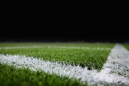 A professional close-up of a green sports field at night, featuring crisp white painted boundary lines on lush turf. The low-angle shot captures the texture of the grass under stadium lighting, with a