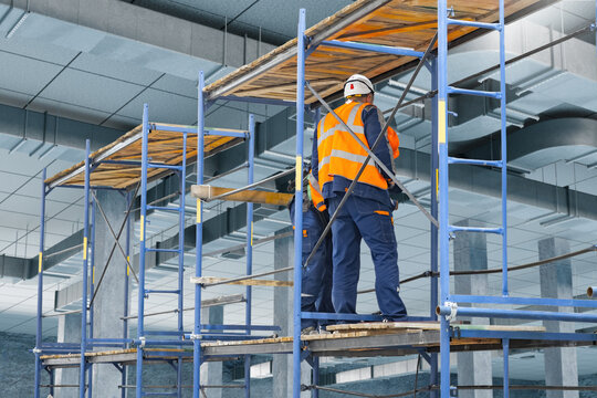 Construction site, Building renovation. Industrial workers in safety gear install ventilation systems using scaffolding inside commercial facility during interior construction works.