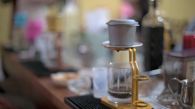 Vietnamese Phin On Shop Counter With Glass Beneath, Blurred Bottles And Tools On Background Shelves, Warm Indoor Lighting, Intimate Cafe Preparation Scene Highlighting Traditional Brewing
