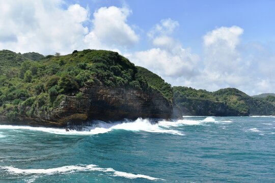 Beautiful view of Timang beach in Yogyakarta, Indonesia with blue sea, crashing waves, green rocky cliffs and cloudy blue sky.