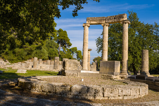Ancient stone columns and circular foundation of the Philippeion stand amid grassy terrain and lush trees in Olympia, Greece. Afternoon sunlight highlights the classical architecture and textured
