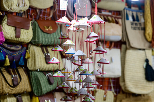 Handcrafted mobiles featuring miniature Vietnamese conical hats hang in front of a display of woven handbags at a market stall in Ho Chi Minh City. The scene highlights artisanal details, colorful