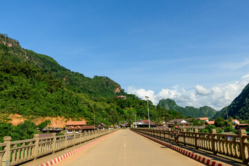 Naklejka premium Wide view of a concrete bridge leading into the village of Nong Khiaw, Laos, surrounded by lush green limestone hills and traditional houses under a clear blue sky. The scene highlights rural