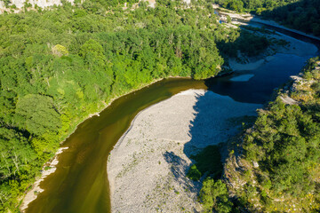 Obraz premium A winding river flows around a wide gravel bar bordered by dense green forest in the Gorges de l'Ardeche. Sunlight casts sharp shadows, highlighting the textures of rocks and foliage.