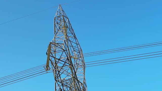 Close view of steel transmission tower with insulators and high voltage power lines cutting across clear blue sky, emphasizing grid infrastructure and industrial design