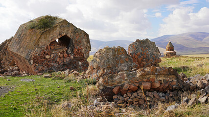 A large, crumbling stone ruin with a hexagonal shape and a central hole stands among scattered rock debris in a grassy field with mountains in the distance at Ani, Turkey © Gregory
