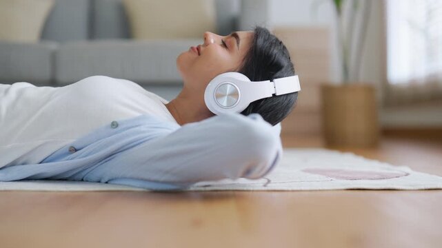 Young Asian woman relaxing at home. Girl in headphones listen to music in cozy living room, using audio healing apps for mental health. Female wellness and self-care lifestyle, ASMR sound therapy