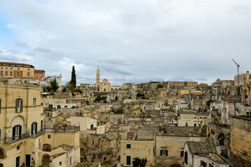 Fototapeta premium Wide view of Matera, Italy, featuring a dense array of historic stone buildings, textured facades, and the prominent cathedral tower rising above the ancient town under an overcast sky.