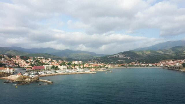 Scenic panoramic view of Banyuls sur Mer with a marina, waterfront buildings, and lush green mountains under a cloudy sky in southern France.