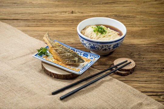 A golden fried fish fillet served on a rectangular plate with a bowl of thin noodles in clear broth beside it.