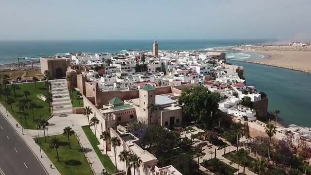 Scenic drone shot capturing the historic Kasbah of the Udayas. Features the fortress walls, traditional white houses, Andalusian gardens, and the Bou Regreg river entering the Atlantic.
