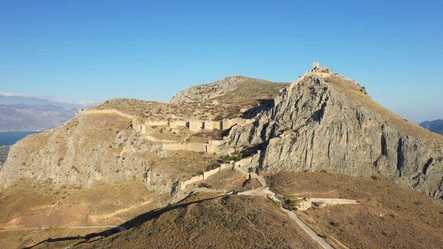 Aerial view of the historic Acrocorinth fortress atop a rugged mountain in the Peloponnese region of Greece under clear blue sky.