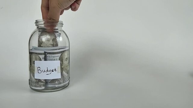 Close-up shot of a hand putting coins or bills into a transparent jar labeled "budget", symbolizing financial planning.