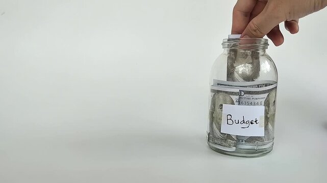 Close-up shot of a hand putting coins or bills into a transparent jar labeled "budget", symbolizing financial planning.