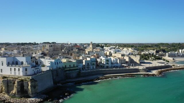 Aerial view of the historic coastal town of Otranto, Italy, featuring traditional Mediterranean architecture along the turquoise shoreline under a clear blue sky.