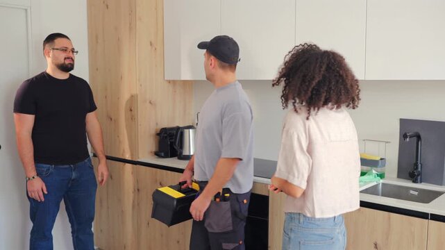 Homeowners speaks with contractor holding toolbox while discussing kitchen renovation progress and cabinet installation details inside modern apartment interior during repair meeting.