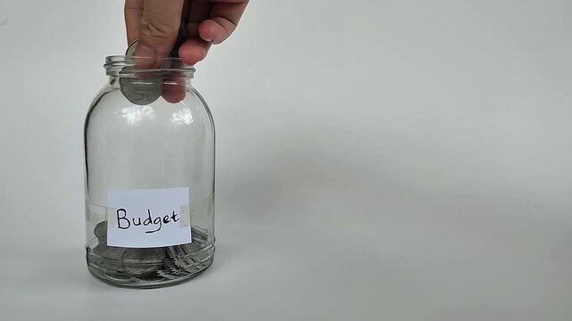 Close-up shot of a hand putting coins or bills into a transparent jar labeled "budget", symbolizing financial planning.